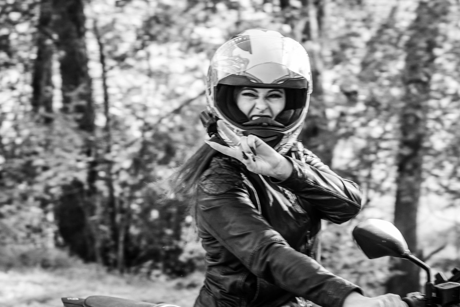 Black and white portrait of a woman in a leather jacket and full-face helmet sitting on a motorcycle in a wooded area.