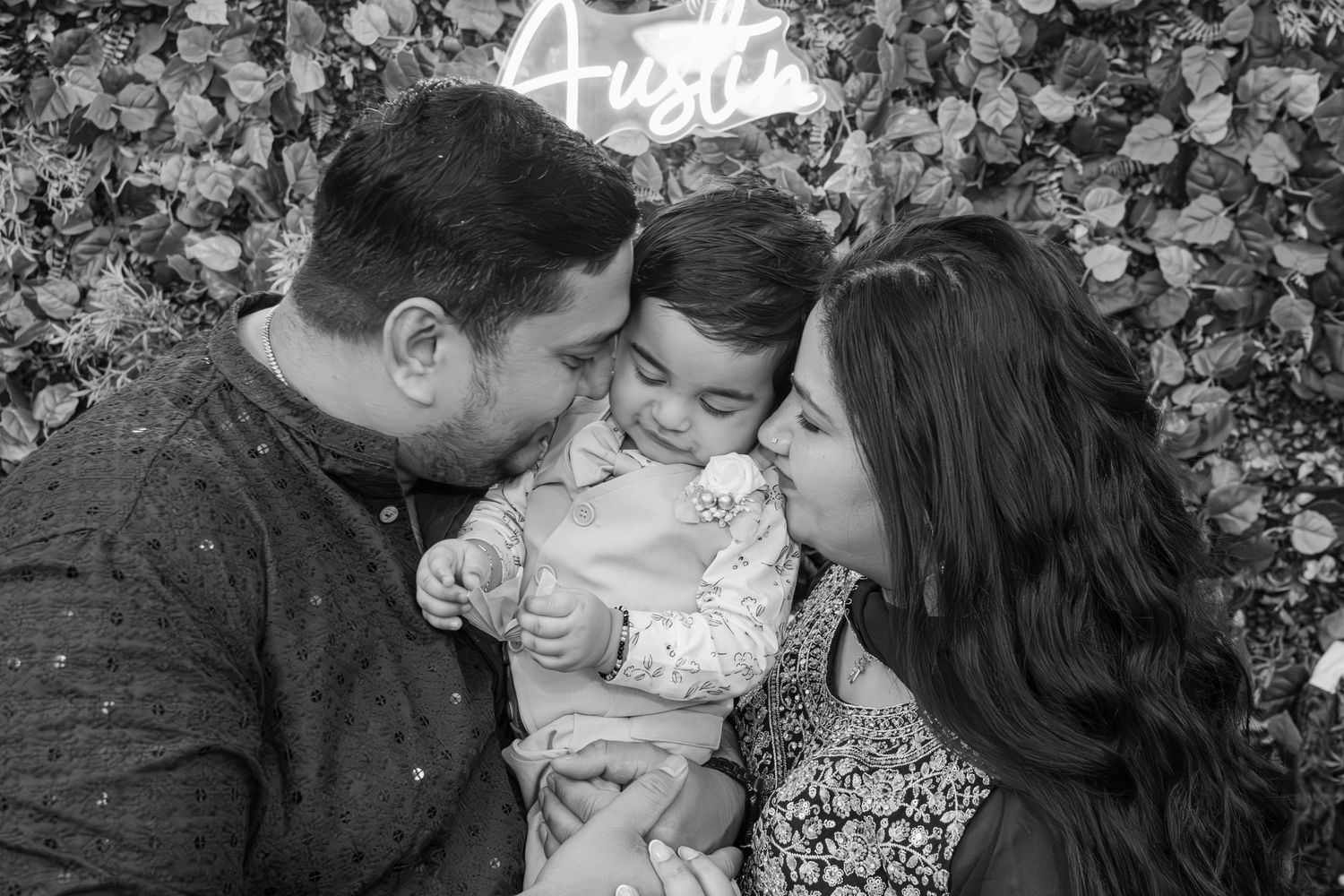 Black and white portrait of parents kissing their toddler son in front of a leafy backdrop with a neon sign at a birthday session.