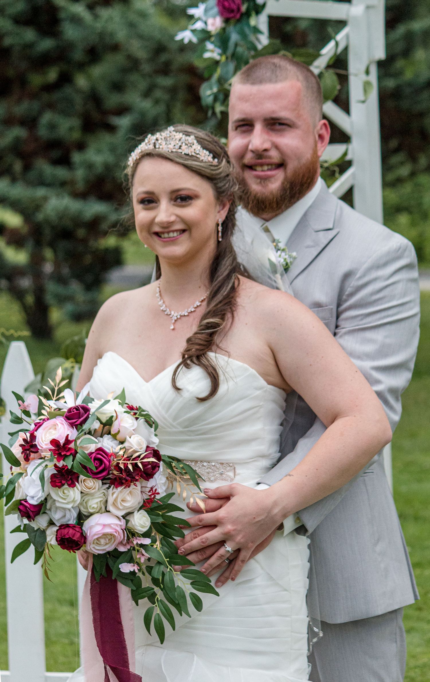 A smiling bride in a tiara holds a burgundy and blush bouquet as her bearded groom in a gray suit embraces her from behind.