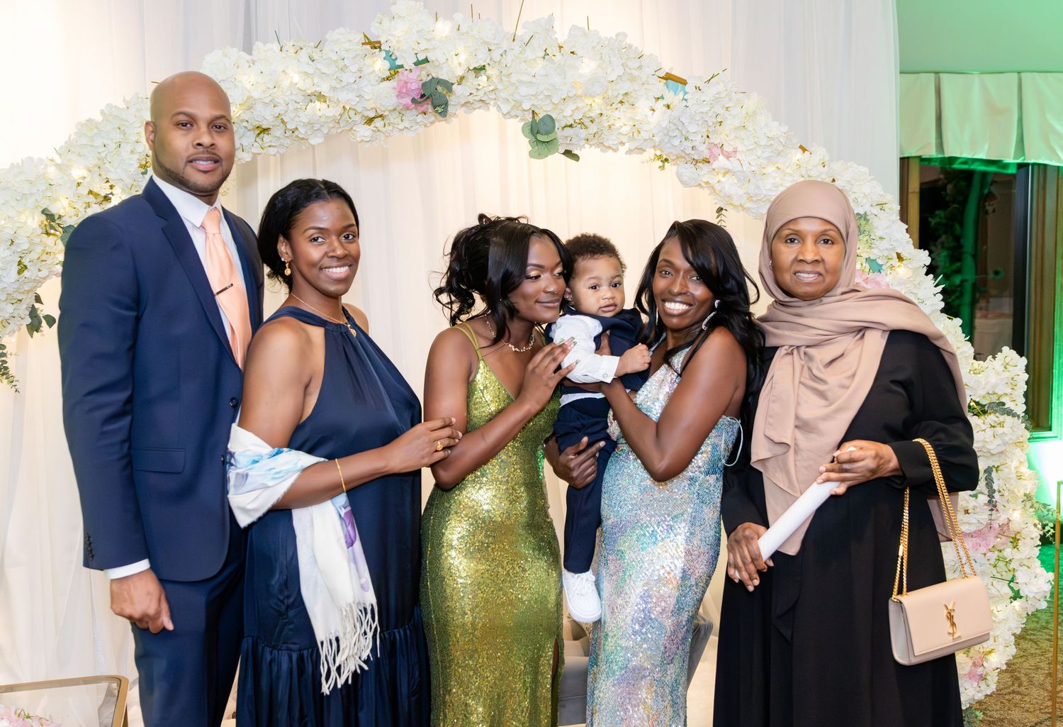 Family of six, including a man in a navy suit, women in elegant dresses and a baby, pose together in front of a floral wedding arch.