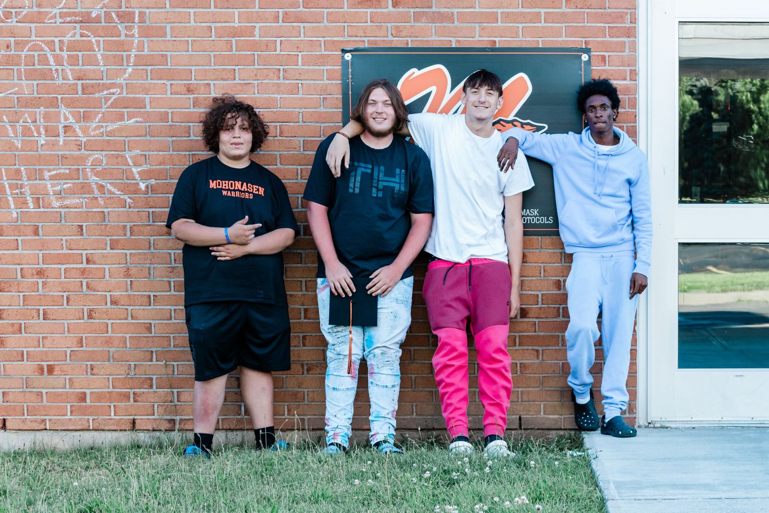 Four teenage friends pose together in front of a red brick school wall with a sign, standing on grass in casual clothes.