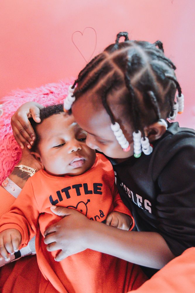 Young girl with beaded braids kisses a sleeping newborn baby in an orange outfit against a pink background with a heart.