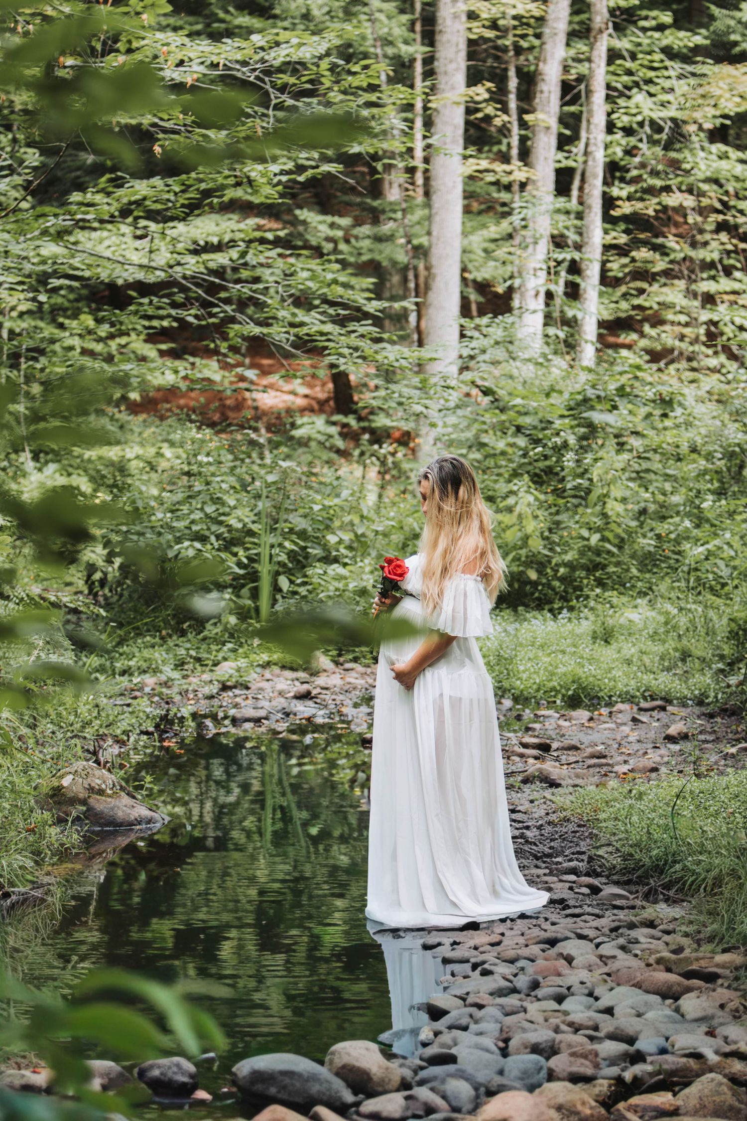 Woman in a flowing white dress holding red flowers stands beside a rocky forest stream.