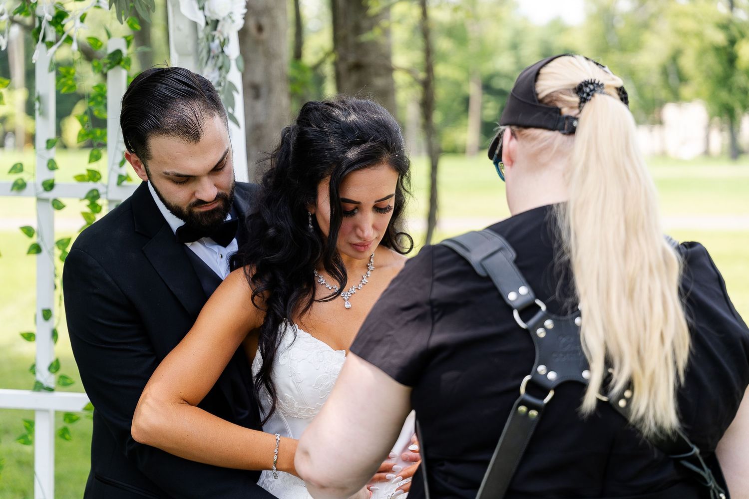 Photographer in a black cap adjusts a bride's dress while the groom in a black tuxedo and bow tie smiles beside her at an outdoor wedding.