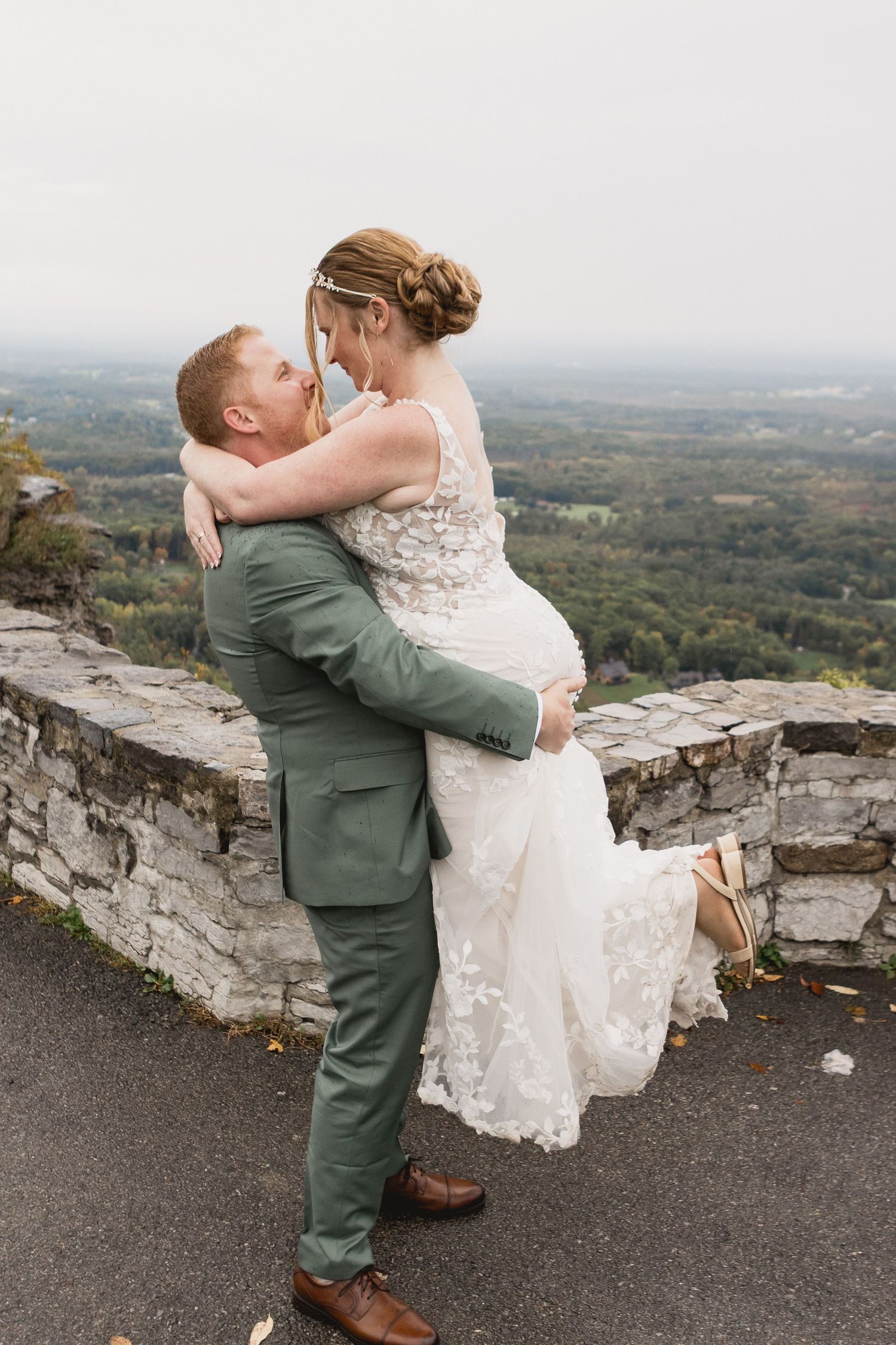 A groom in a green suit lifts his smiling bride in a lace wedding dress on a stone wall overlooking a scenic mountain valley.