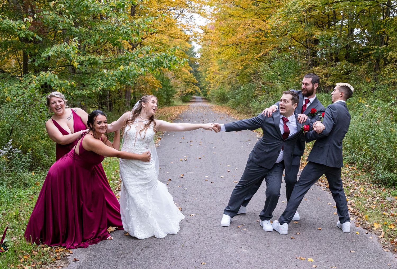 A bride holds hands with her groom as bridesmaids in burgundy gowns and groomsmen playfully tug in a tug-of-war on an autumn path.