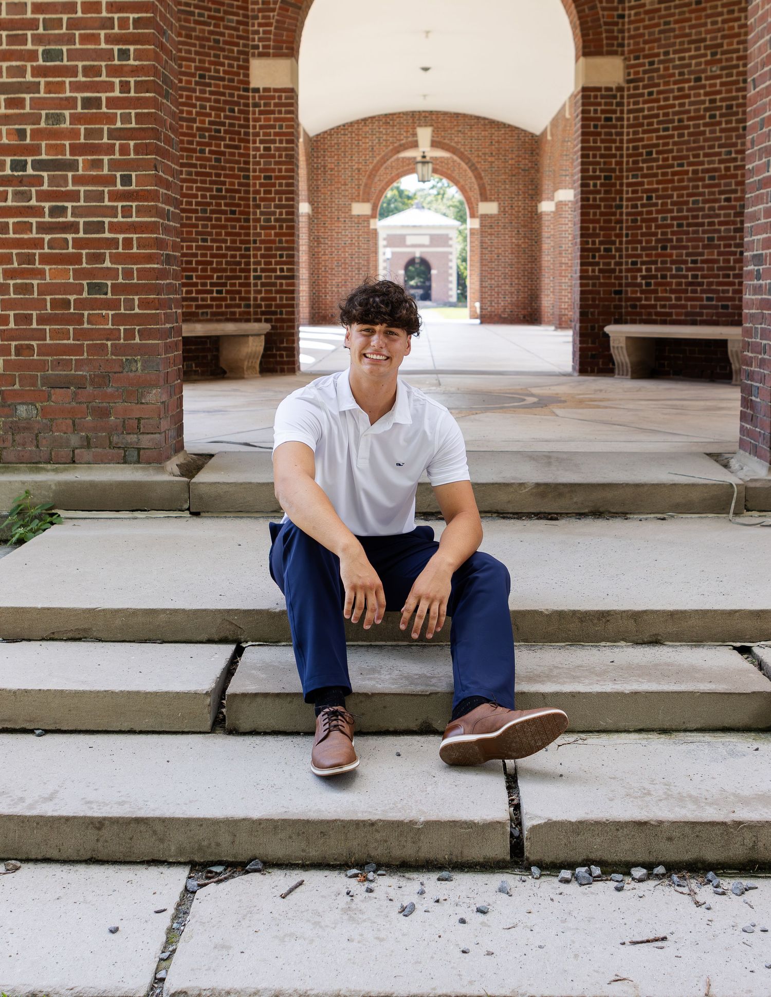 Young man in a white polo and navy pants sits on wide stone steps beneath a red brick arched walkway.