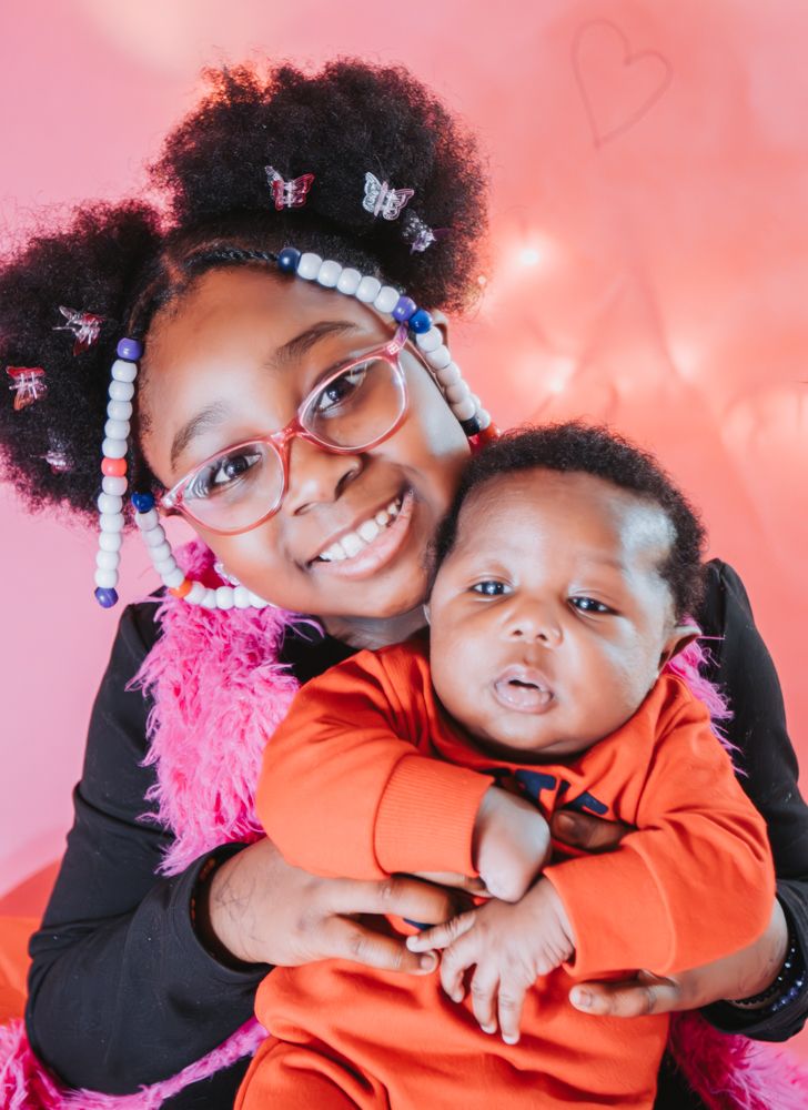 Smiling girl in pink glasses and beaded hair holds a newborn baby in an orange outfit against a pink heart backdrop.