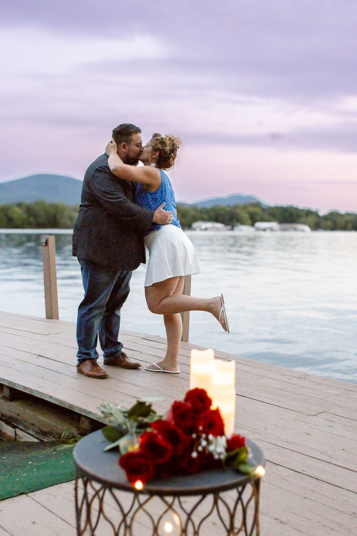 A couple kisses on a wooden lake dock at dusk, with red roses and glowing candles on a table beside them after a proposal.