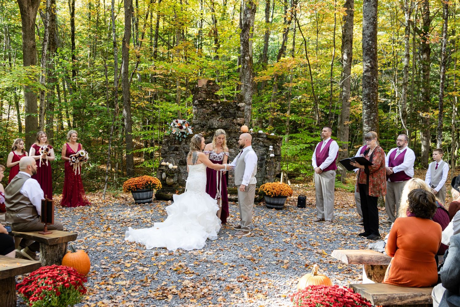 Outdoor autumn forest wedding ceremony with a bride in a white gown, bridesmaids in burgundy, guests seated on benches, and pumpkins nearby.