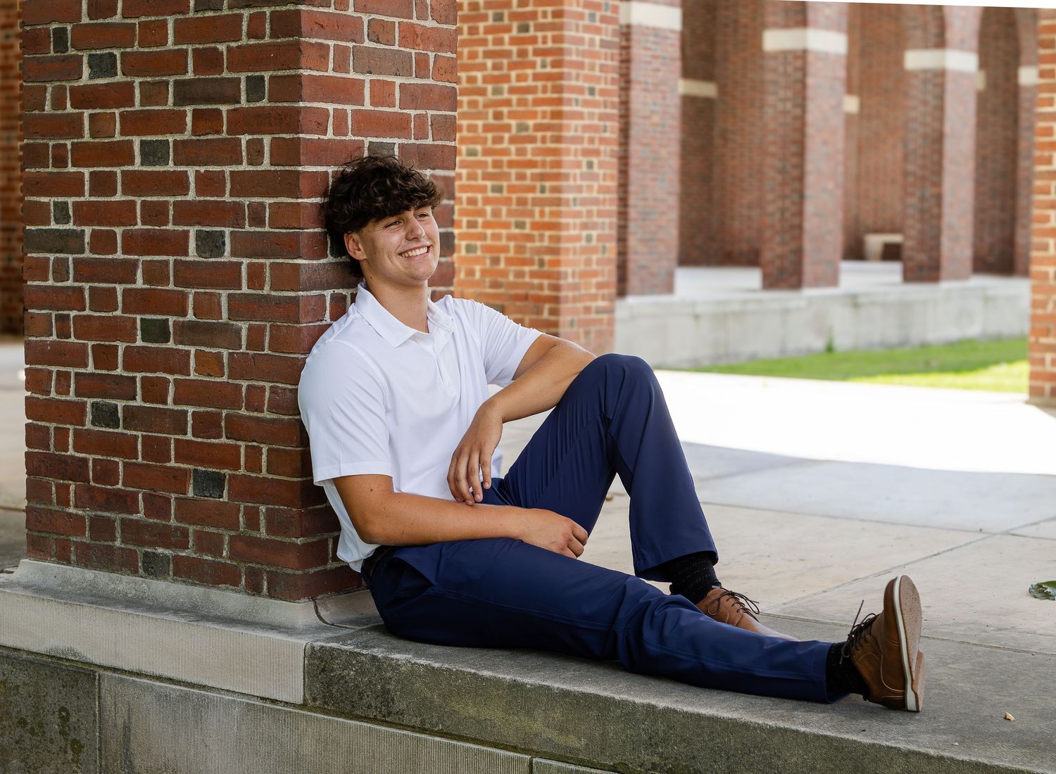 Smiling young man in a white polo and navy pants sits on a stone ledge leaning against a red brick wall.