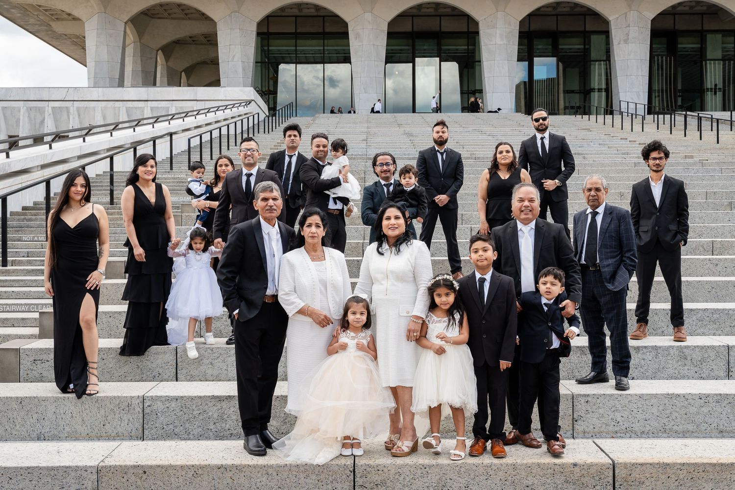 Large multigenerational family in black, white and formal attire posing together on wide stone steps outside a modern building.
