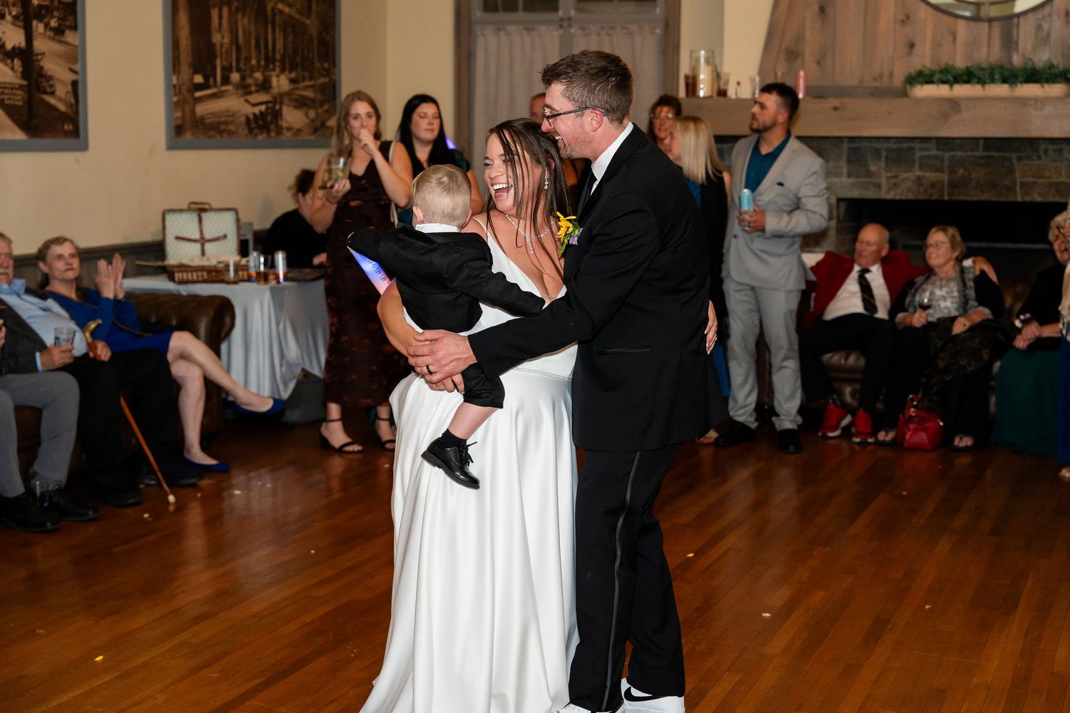 A groom in a black suit dances with his bride in a white gown while holding a small child, as wedding guests watch.
