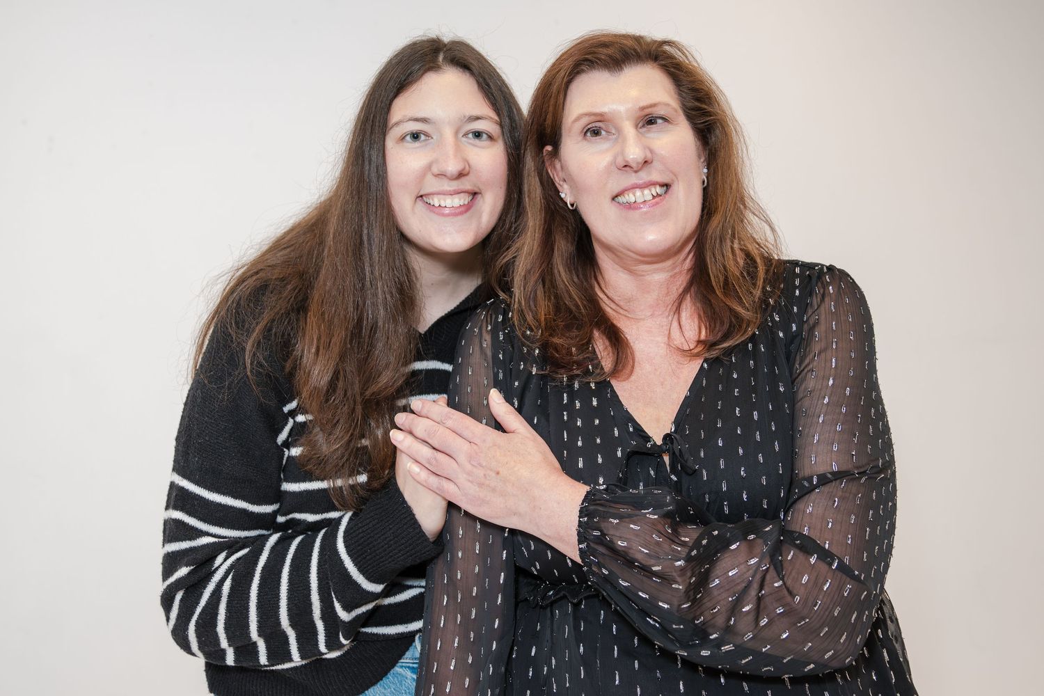 A smiling young woman and her mother embrace in a bright studio portrait, both wearing black patterned tops.