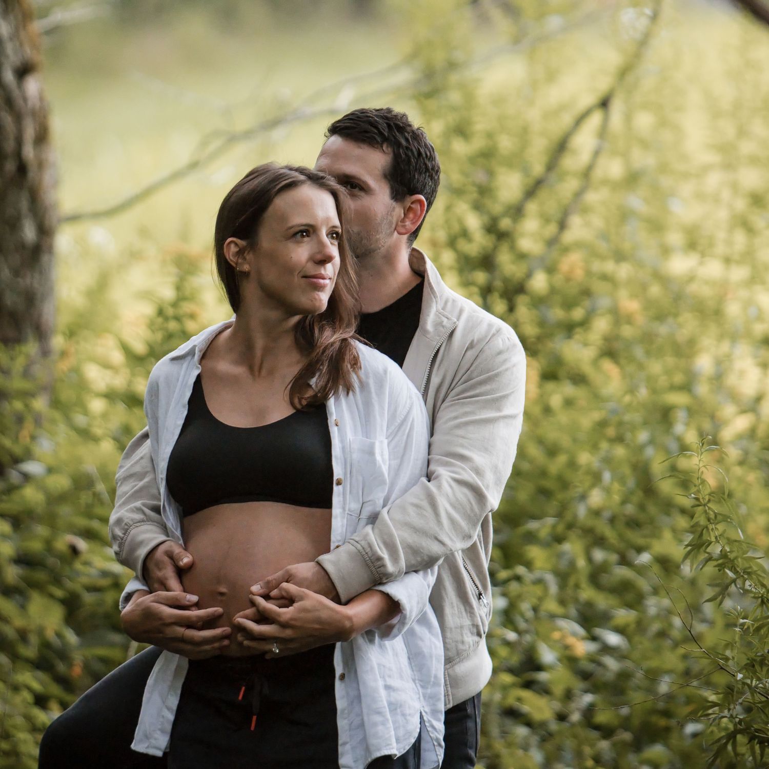 A man embraces his pregnant partner from behind in a green forest, both hands resting on her bare baby bump.