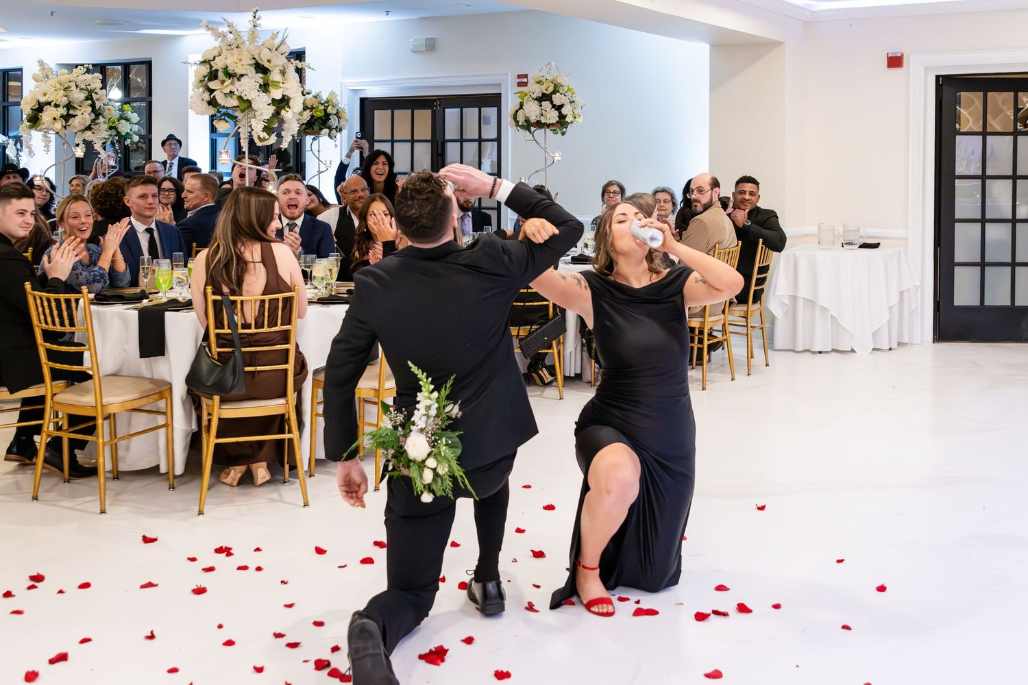 Groom in a black suit dances with a bride in a black dress on a reception dance floor scattered with red rose petals as guests watch.