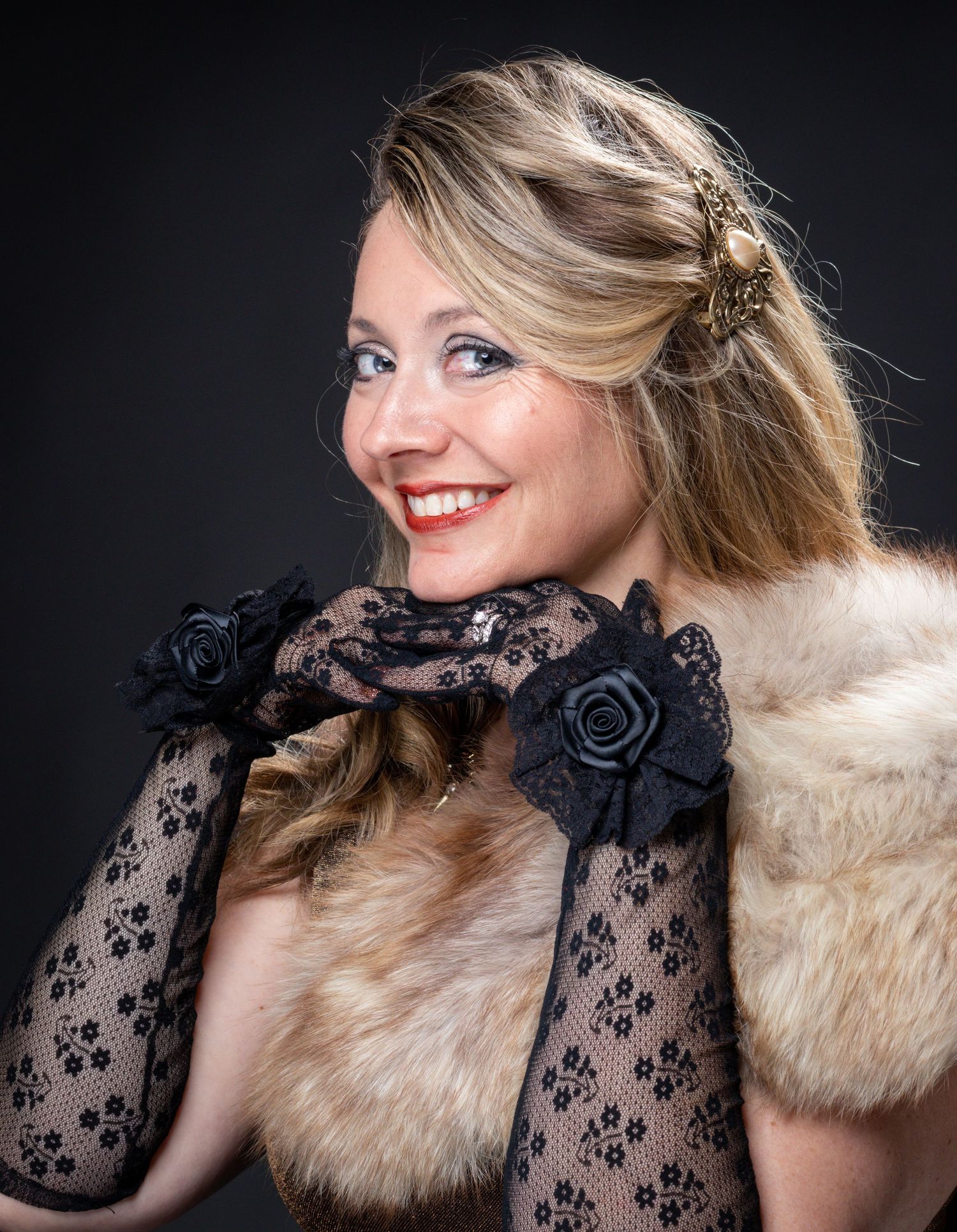 Smiling woman with blonde hair wearing black lace gloves with rose accents and a vintage fur stole in a studio portrait.
