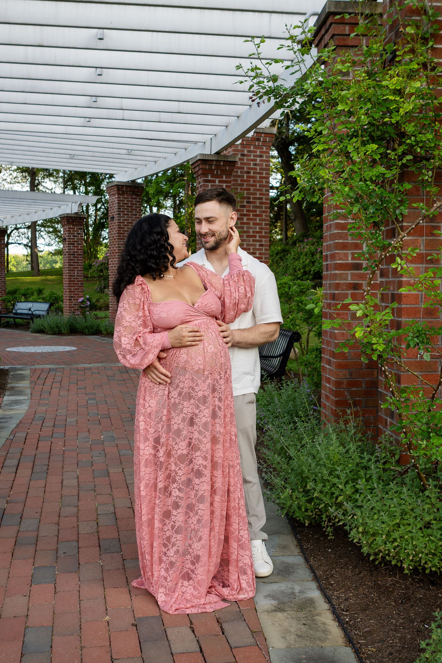 A couple in a pink lace maternity gown and white shirt embrace under a brick pergola in a garden.