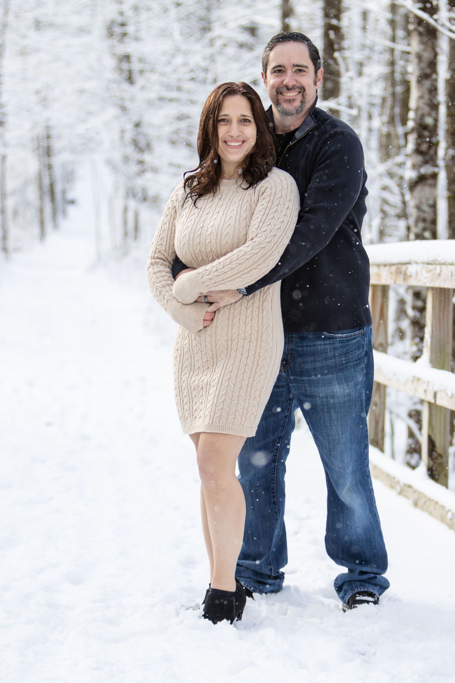 Smiling couple embraces on a snowy path, the woman in a beige knit sweater dress and the man in a dark sweater and jeans.