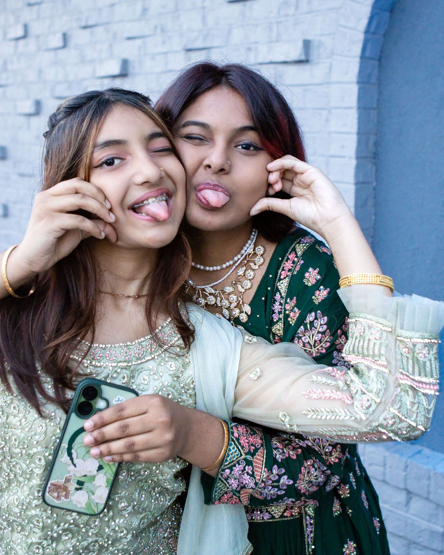 Two smiling young women in embellished green traditional outfits playfully stick out their tongues while posing against a blue brick wall.