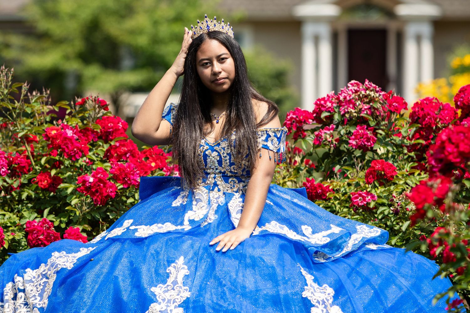 Young woman in a sparkling royal blue quinceañera gown and tiara posing gracefully among blooming red rose bushes in a sunny garden.