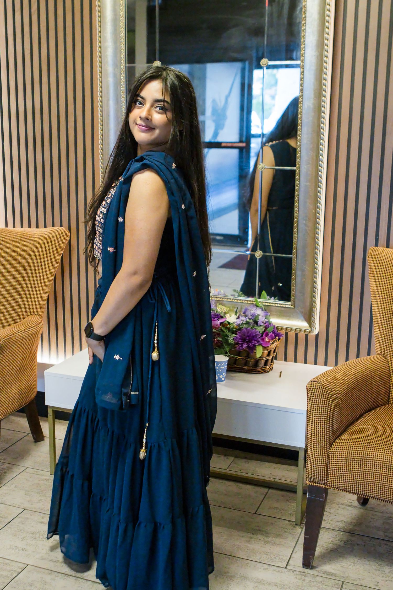 Smiling woman in a flowing navy blue ruffled dress with dupatta stands beside a white table with purple flowers and a gold-framed mirror.