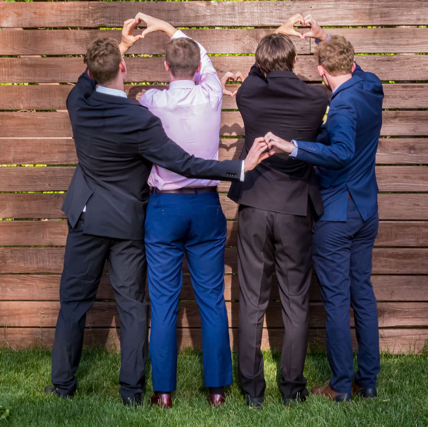Four men in suits stand with their backs to the camera, arms linked, making heart shapes with their hands in front of a wooden fence.