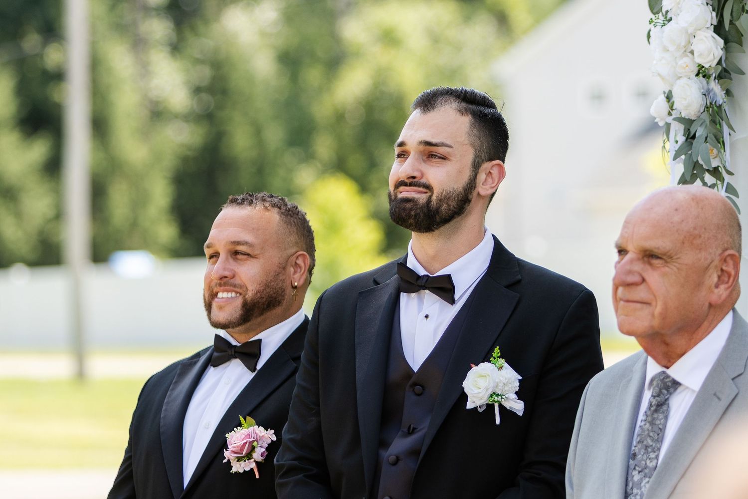 Three men in formal wear, two with boutonnieres, watch a wedding ceremony outdoors with white flowers hanging nearby.