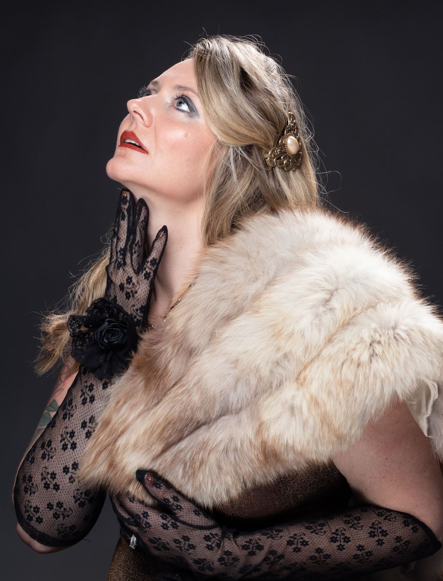 Woman with wavy blonde hair wears a fur stole and black lace gloves while gazing upward in a dramatic studio portrait.