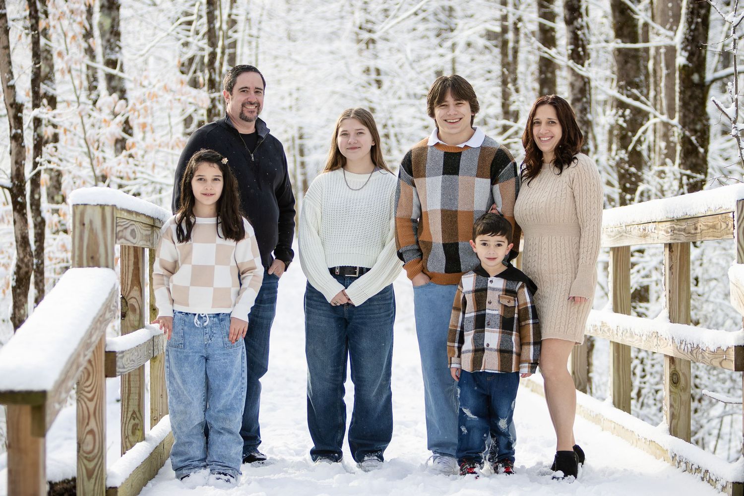 Family of six in cozy winter clothes stands on a snow-covered wooden bridge surrounded by frosty trees in a winter forest.