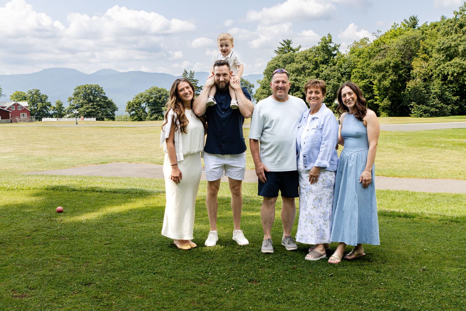 A smiling multi-generational family poses on a grassy field with mountains behind them, a man holding a toddler on his shoulders.