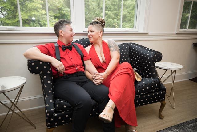 Two brides in matching red outfits sit on a polka-dot sofa holding hands and smiling at each other beside a bright window.