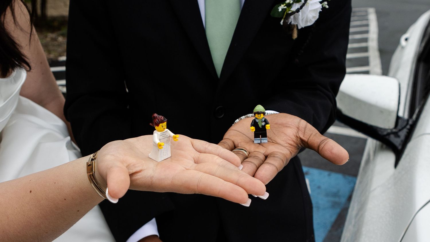 A couple holds tiny bride and groom LEGO figurines in their open palms during their wedding day.