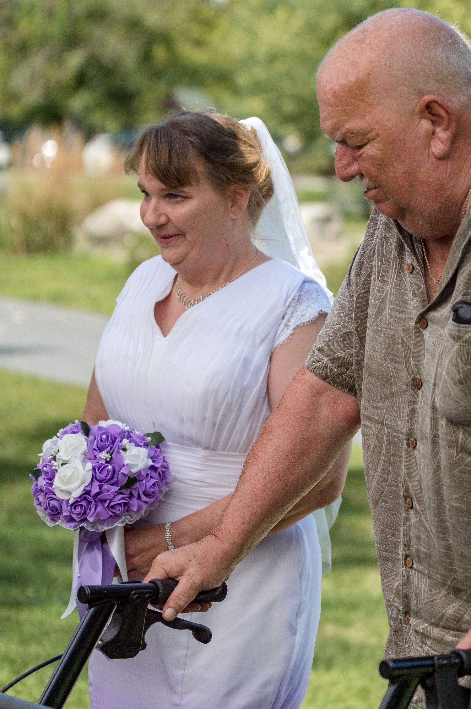 A bride in a white dress and veil holding a purple and white bouquet walks outdoors with an older man steadying a walker.