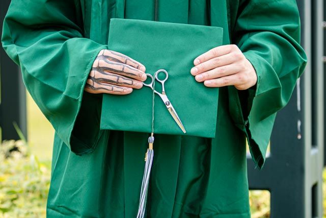 Hands with skeleton tattoos hold a green graduation cap and silver hair shears, with a tassel hanging beside the green gown.