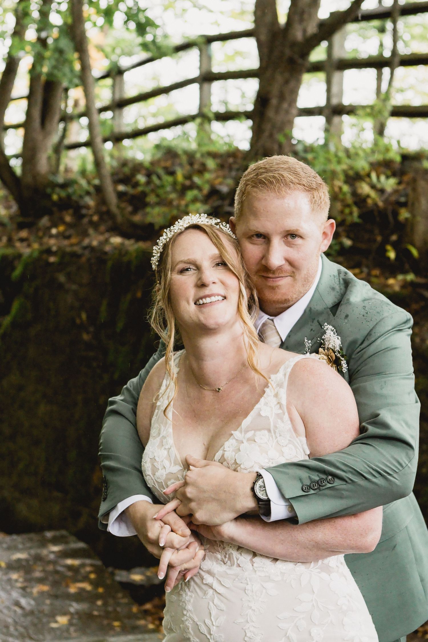 Smiling bride in a lace gown and floral hair crown is embraced from behind by her groom in a green suit in a wooded outdoor setting.