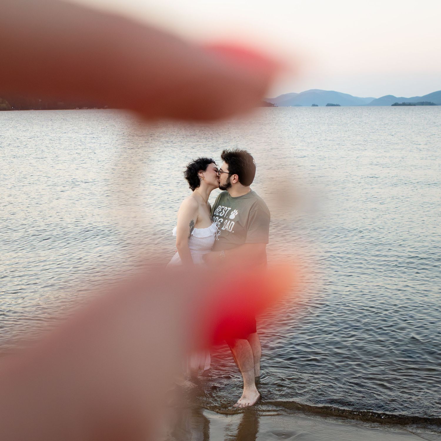 Couple kissing in shallow lake water at sunset, framed by a blurred pink fingertip in the foreground.