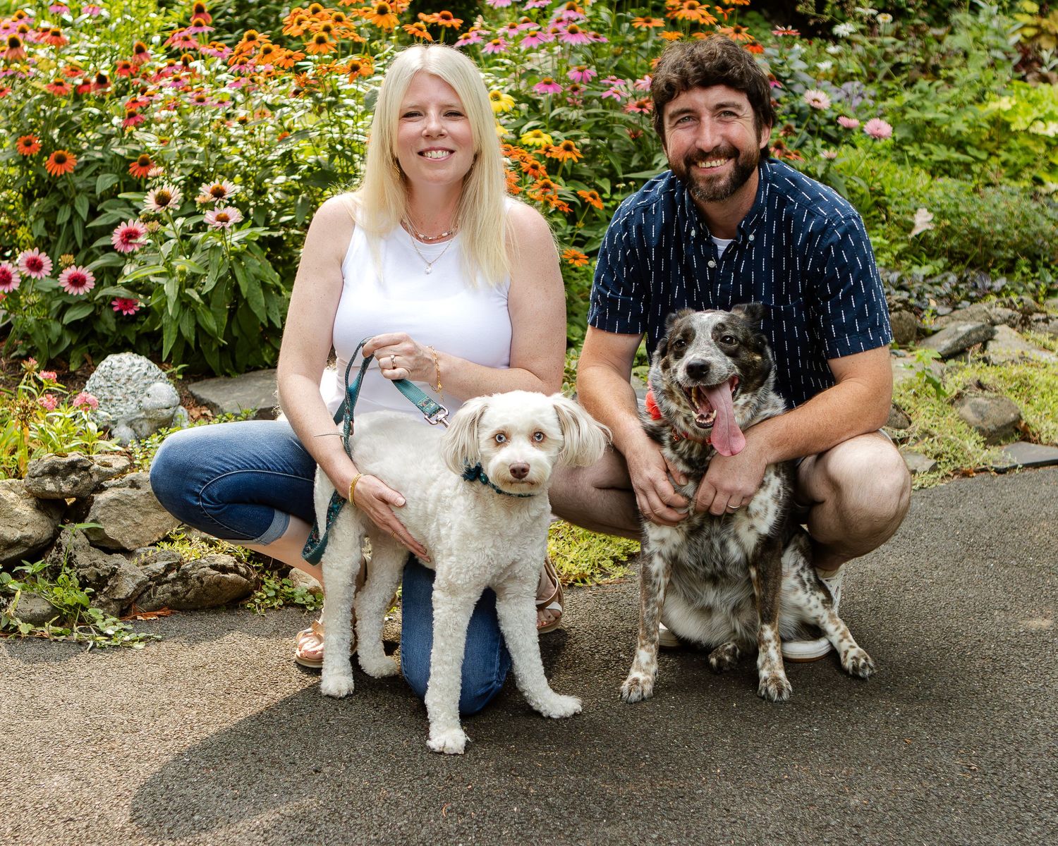 A smiling woman and bearded man crouch outdoors with their two dogs in front of colorful orange and pink flowers.