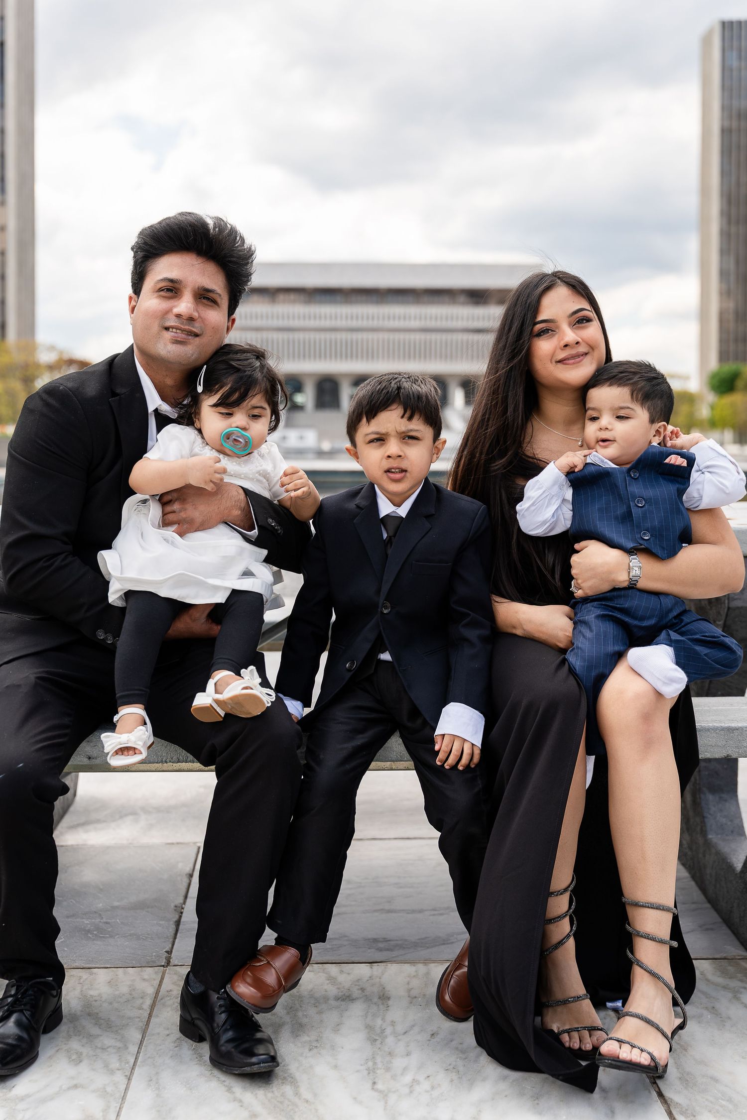 A family of four in formal black attire sits together on a stone bench with a modern building behind them.