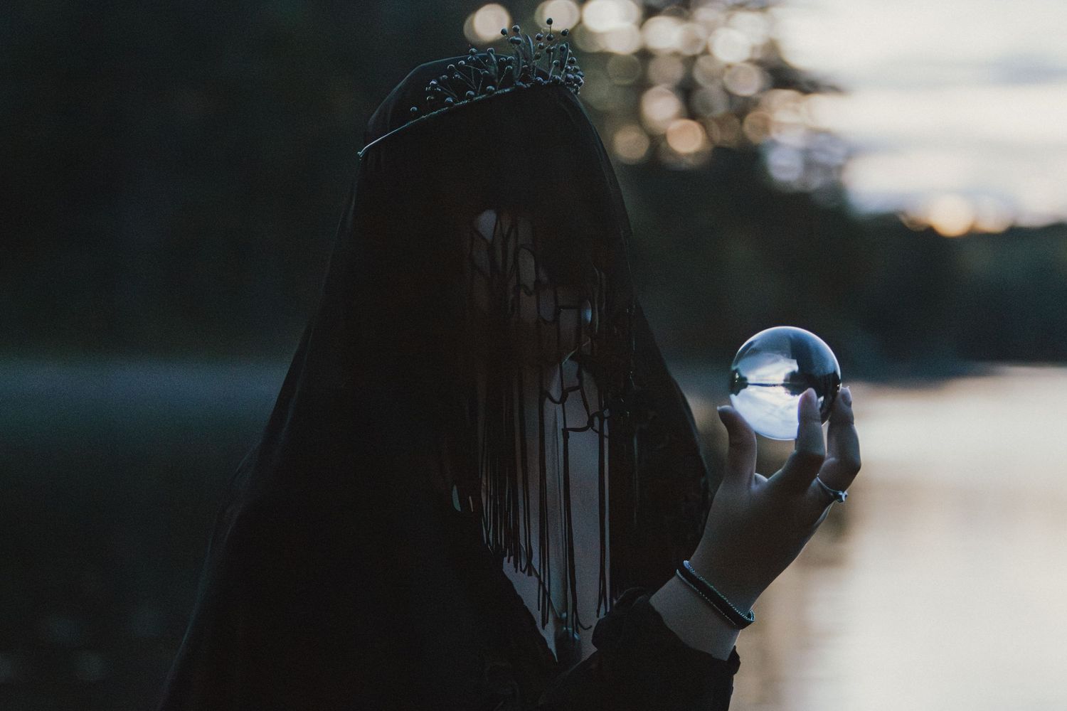 A veiled figure in a black hooded cloak and dark crown holds a glass crystal ball beside a misty lake at dusk.