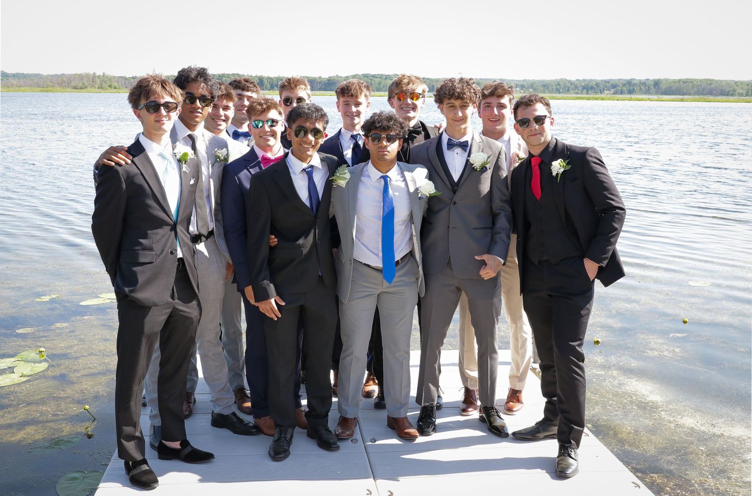 Group of young men in suits and boutonnieres posing together on a wooden dock beside a calm lake on a sunny day.