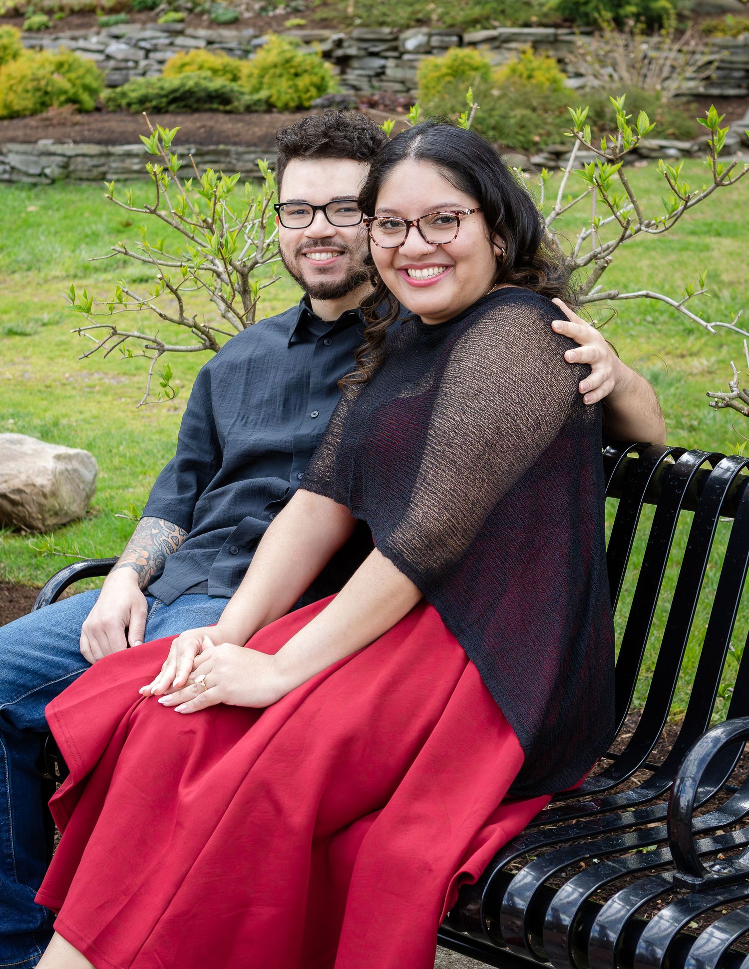 A happy engaged couple sits together on a black garden bench, with the woman in a red skirt showing her engagement ring.