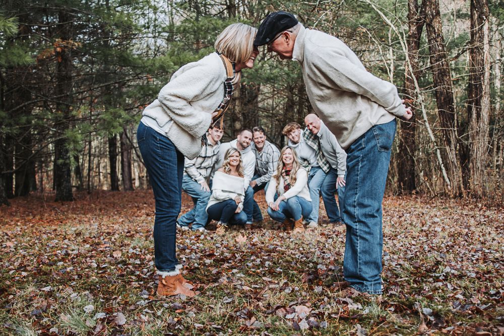 Multigenerational family in cozy sweaters gathers around a baby on autumn leaves in the woods.