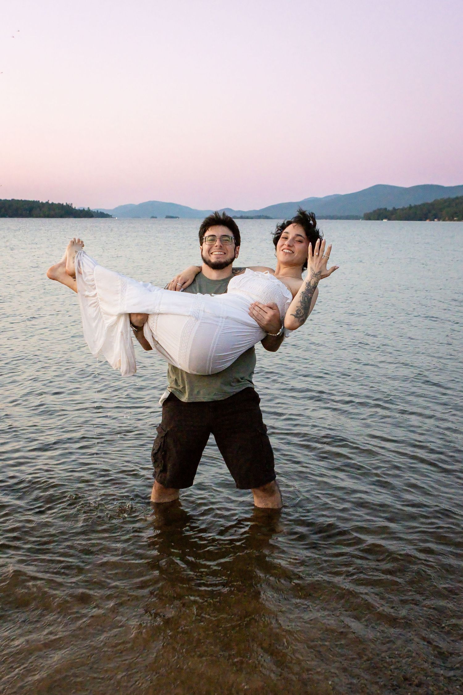 Man standing in shallow lake water carries a laughing woman in a white dress during a sunset engagement photo shoot.