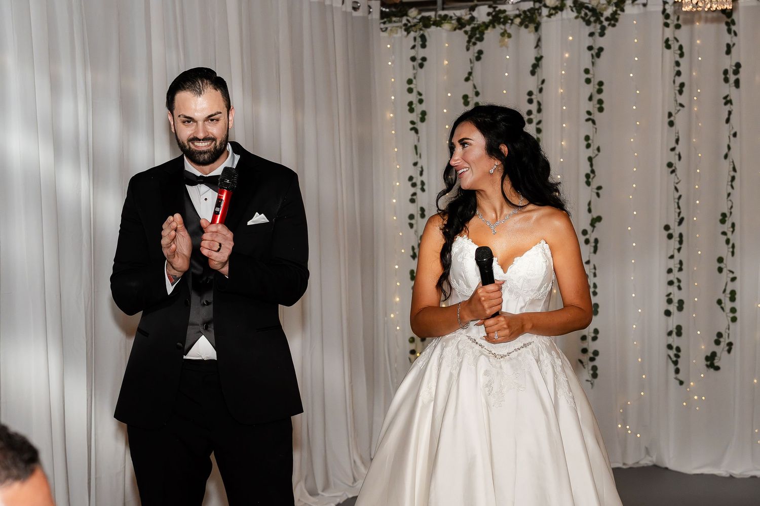 A groom in a black tuxedo applauds while a bride in a white gown holds a microphone during a wedding reception speech.