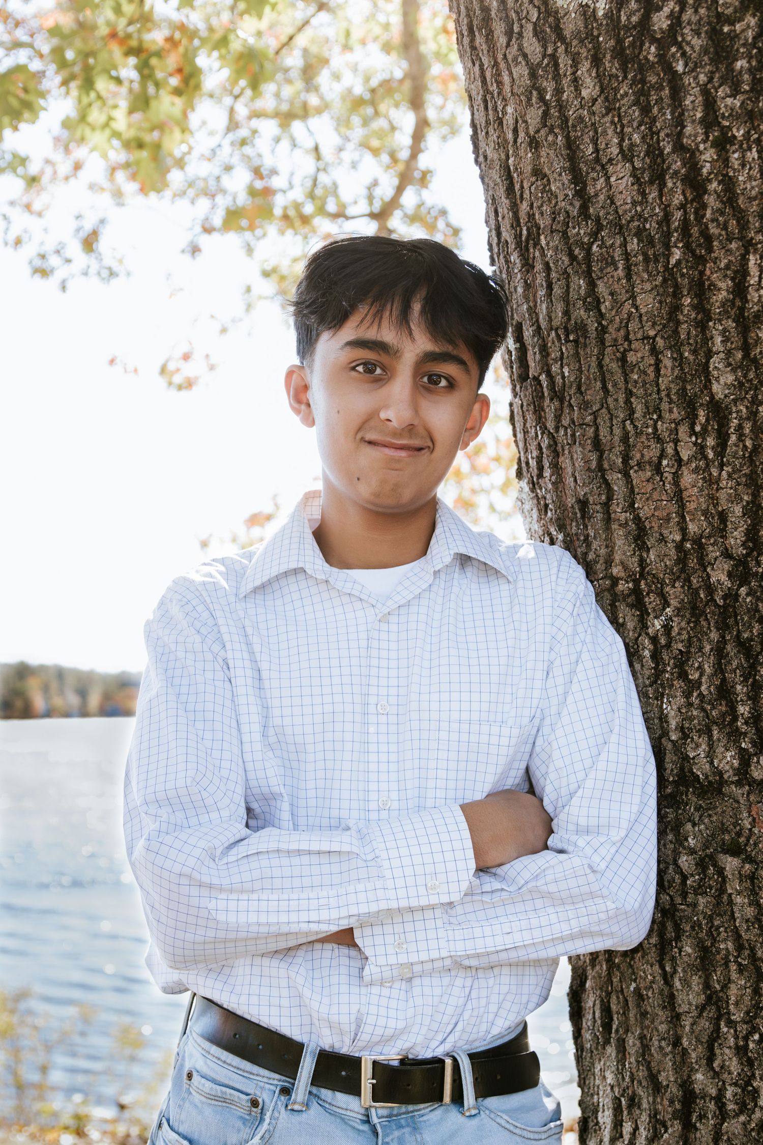 A teen in a light checkered button-up shirt leans against a tree with arms crossed, with a calm lake in the background.