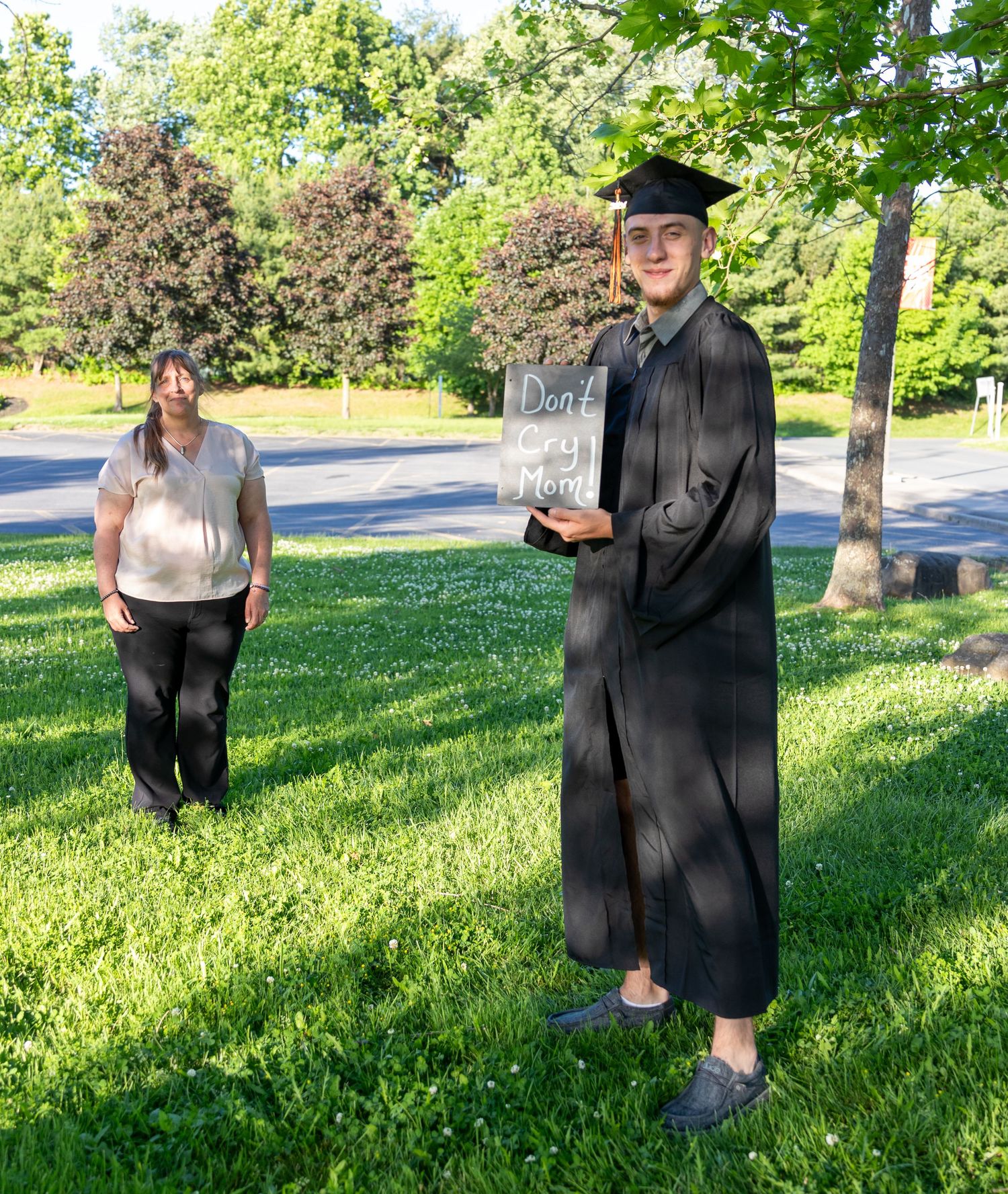 A graduate in a cap and gown holds a chalkboard sign reading Don't Cry Mom while a woman stands on the grass behind him.