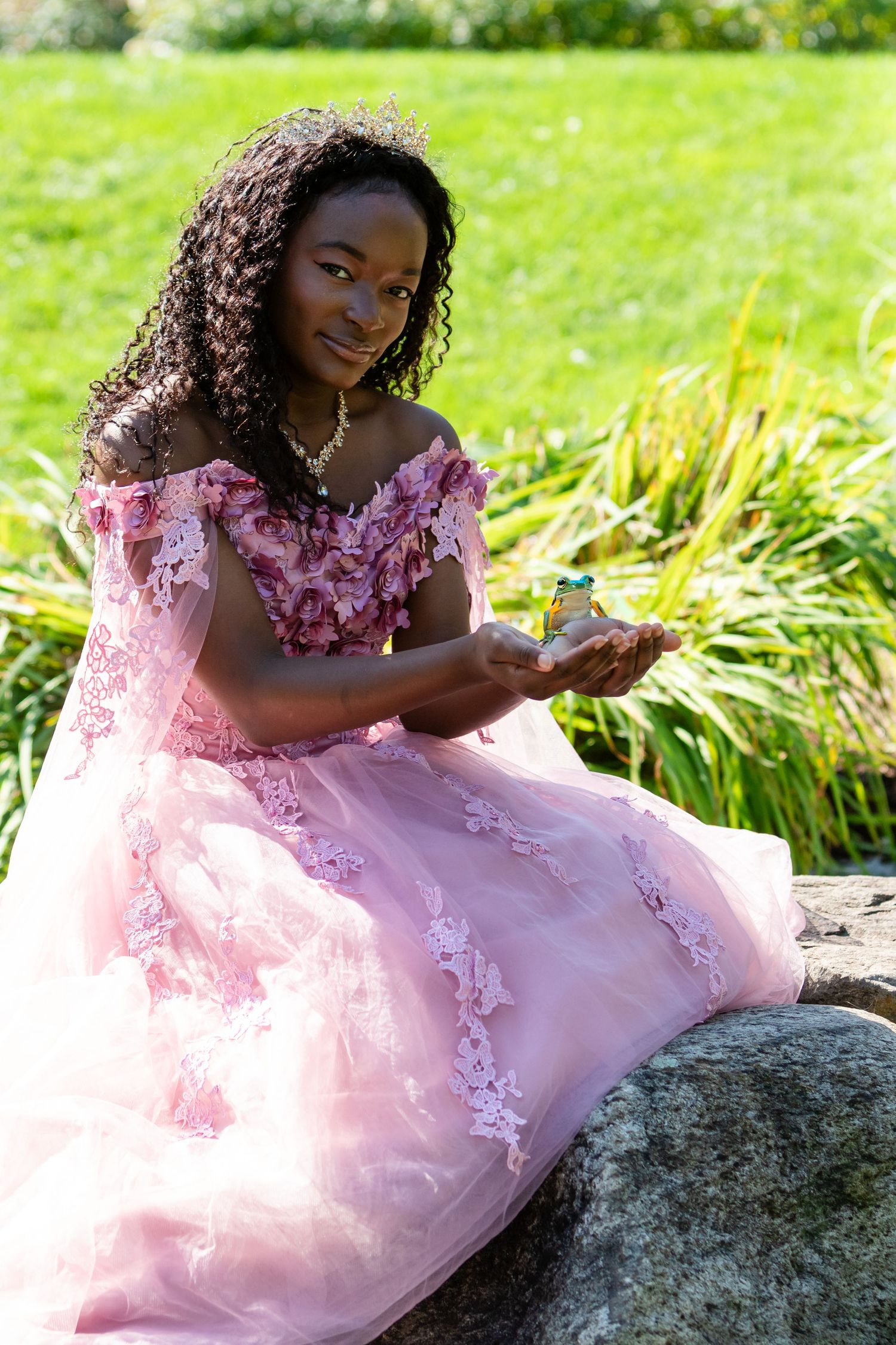 Young woman in a pink floral tulle gown sits on a large rock holding a small item in her hands beside green grass.