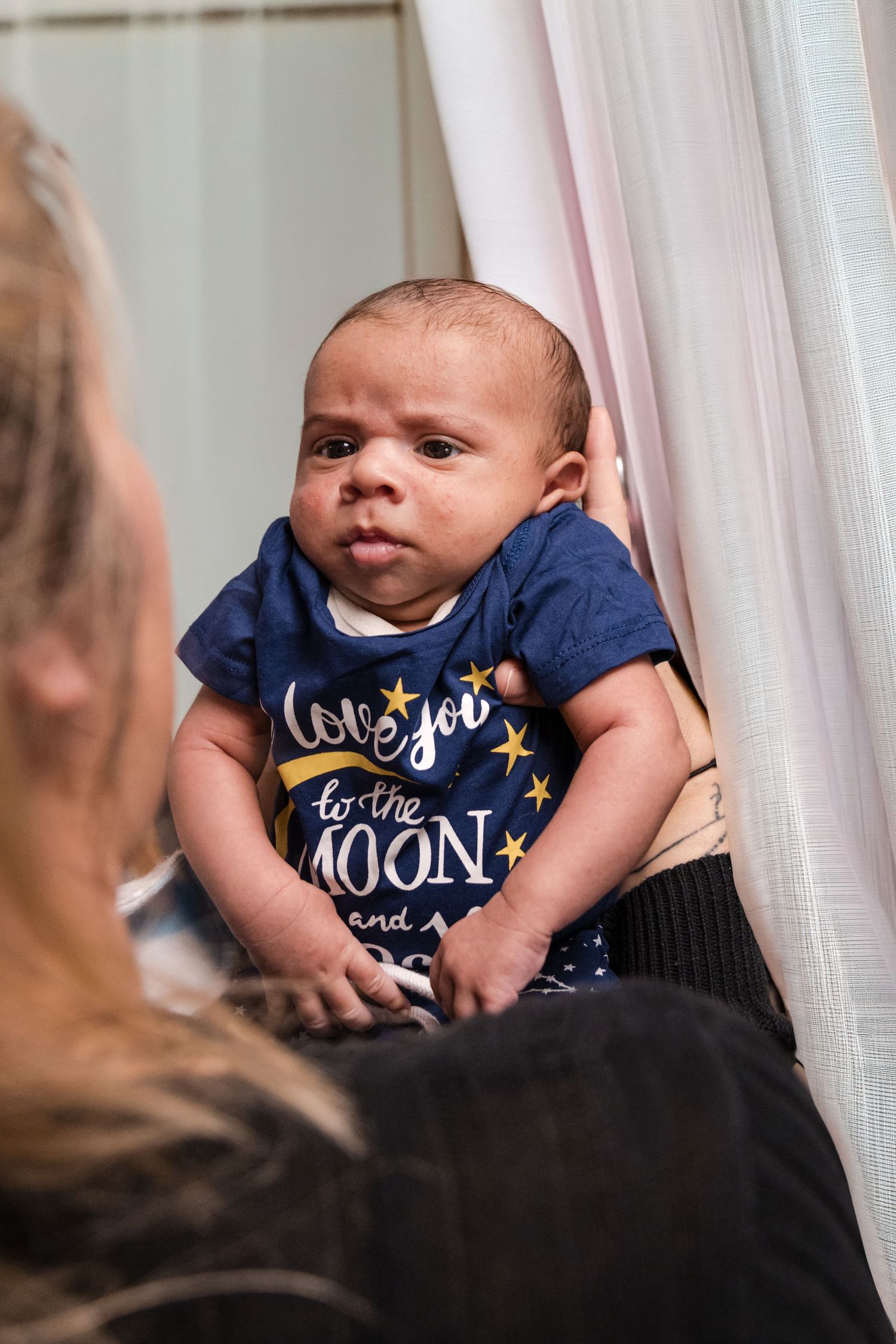A wide-eyed newborn in a navy love you to the moon onesie is held up by a woman in front of sheer white curtains.