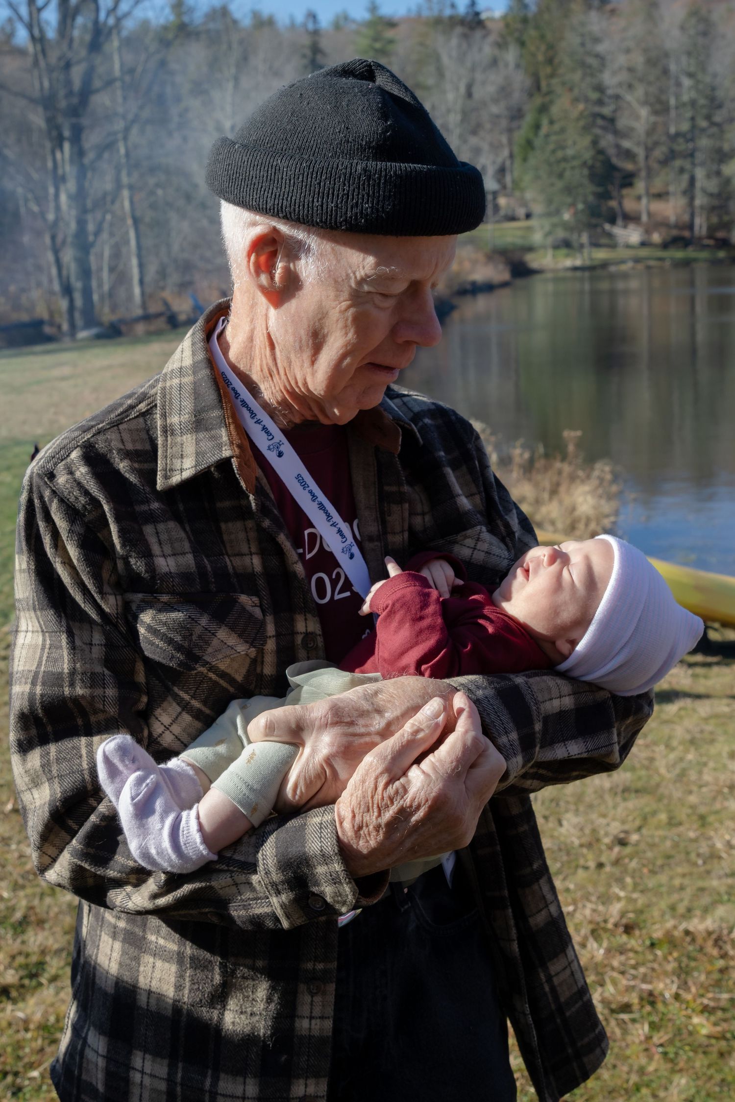 An elderly man in a black knit cap and plaid jacket tenderly cradles a sleeping baby beside a lake.